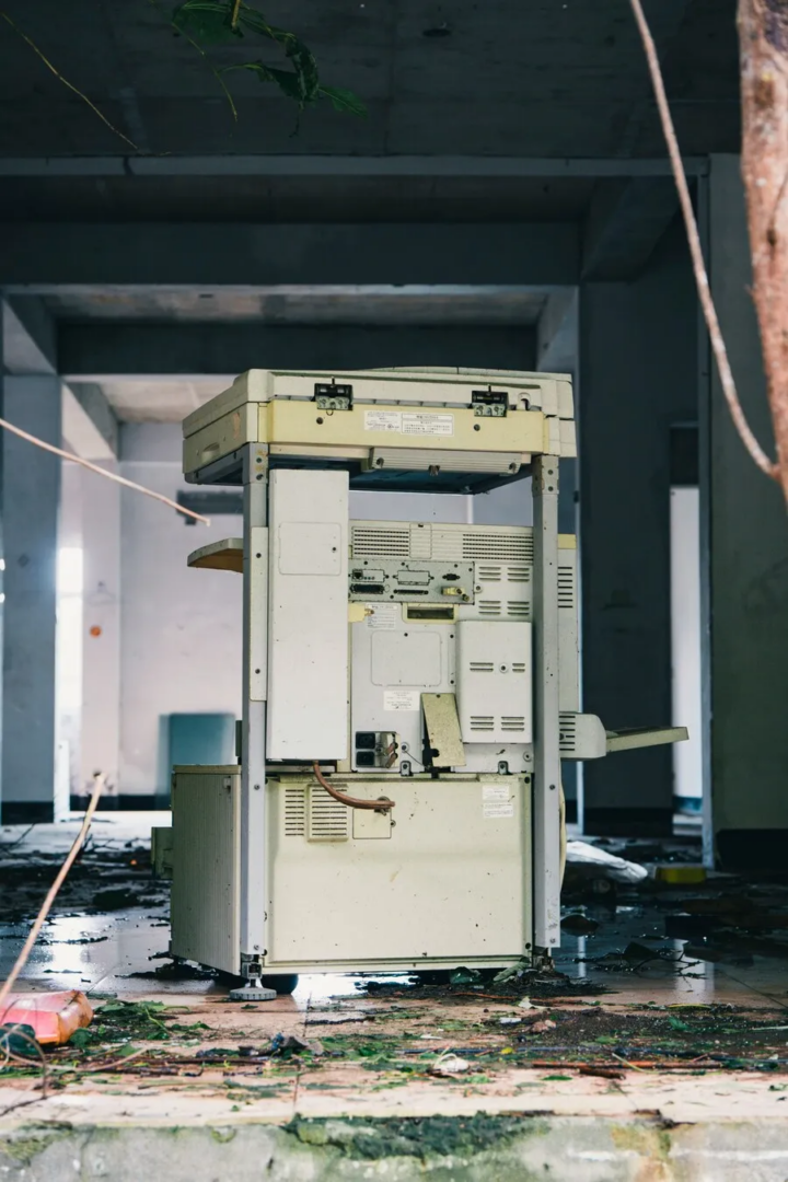 Discarded machine standing inside an abandoned building, surrounded by debris, exposed wiring and vegetation.