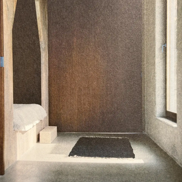 Minimalist interior with textured brown walls, a low white built-in bench, dark rectangular rug and tall window admitting natural light.
