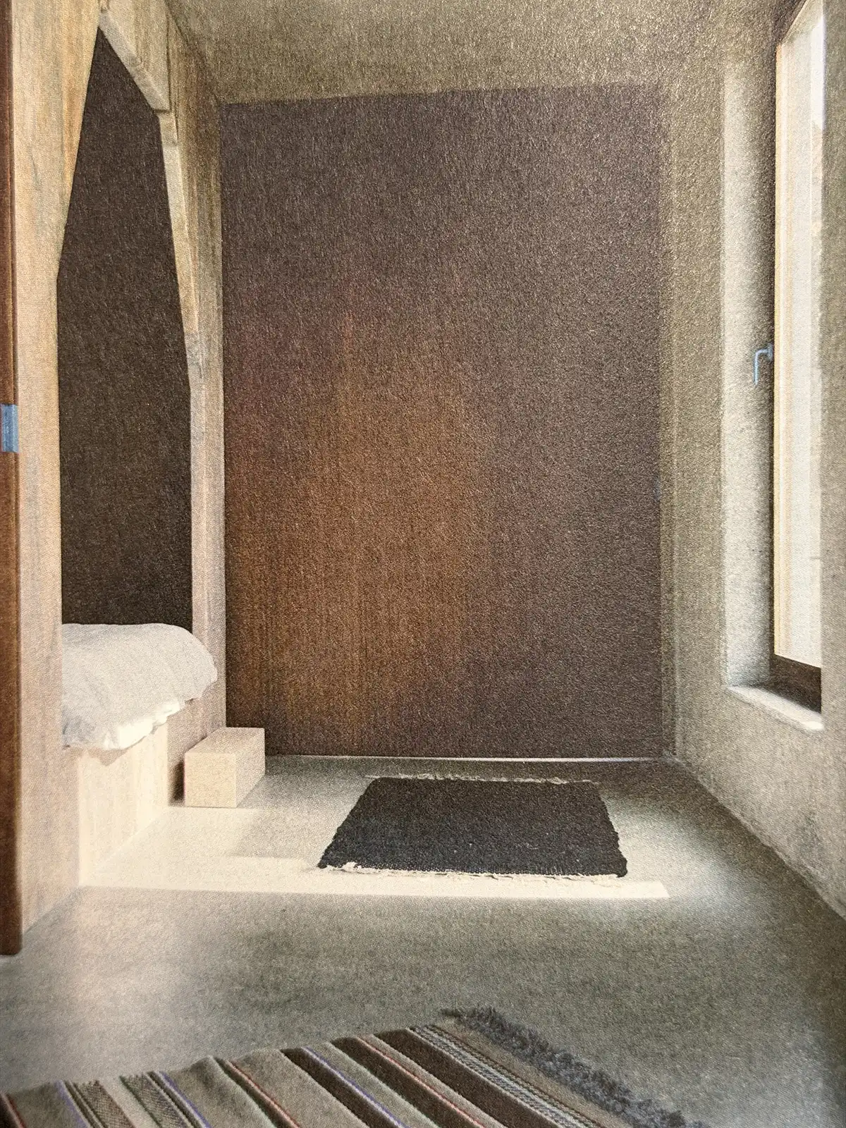 Minimalist interior with textured brown walls, a low white built-in bench, dark rectangular rug and tall window admitting natural light.