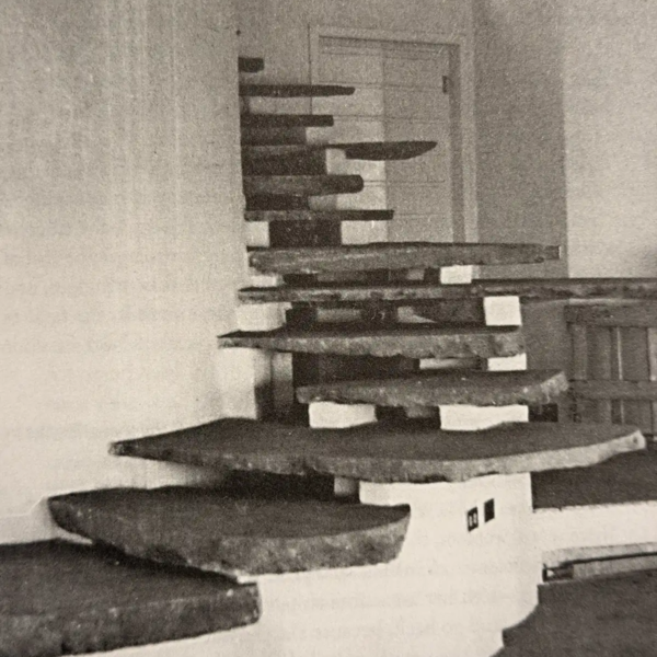 Black-and-white architectural photograph showing a sculptural staircase composed of projecting, irregularly shaped steps arranged around a central vertical support.