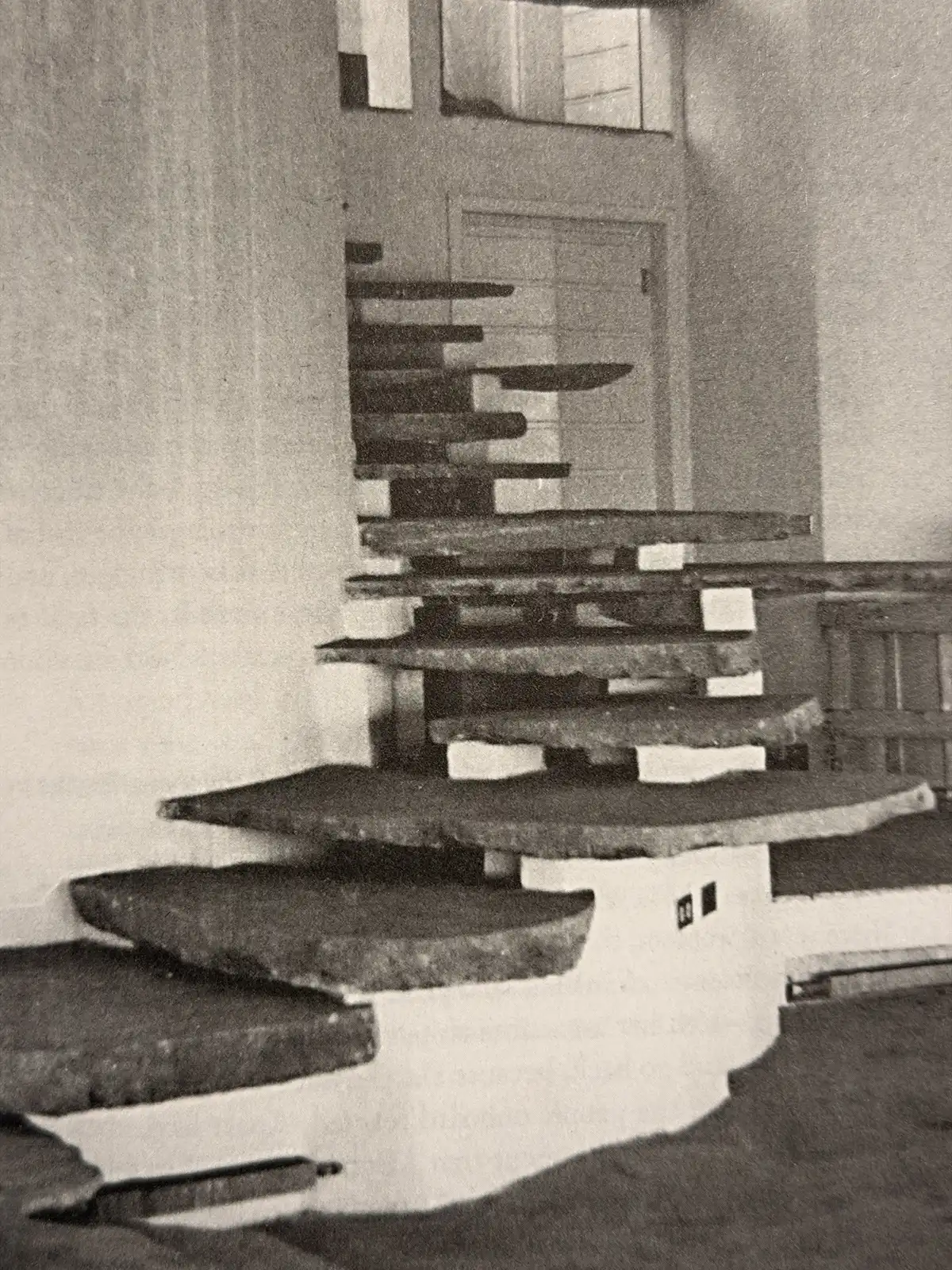 Black-and-white architectural photograph showing a sculptural staircase composed of projecting, irregularly shaped steps arranged around a central vertical support.